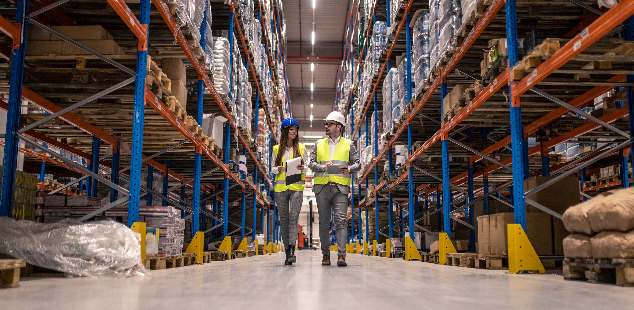 Dois supervisores de logística, um homem e uma mulher, vestindo coletes refletivos e capacetes de segurança, caminham pelo corredor central de um armazém industrial moderno. Eles conversam enquanto seguram uma prancheta e um tablet, ladeados por estantes altas repletas de mercadorias paletizadas.