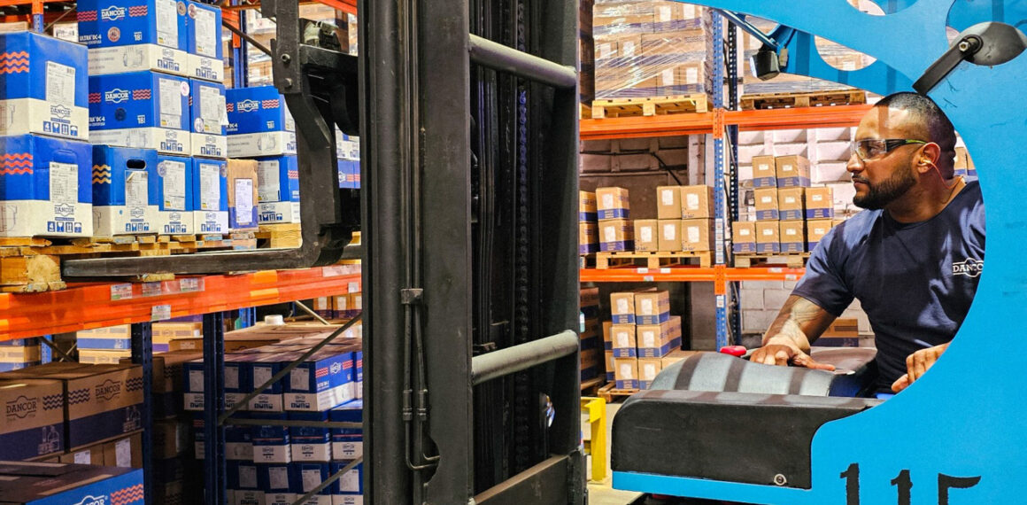 Operador de empilhadeira em um armazém logístico organizado, utilizando equipamentos de segurança e operando a máquina para movimentar pallets de caixas de produtos Dancor em prateleiras industriais de alta densidade.