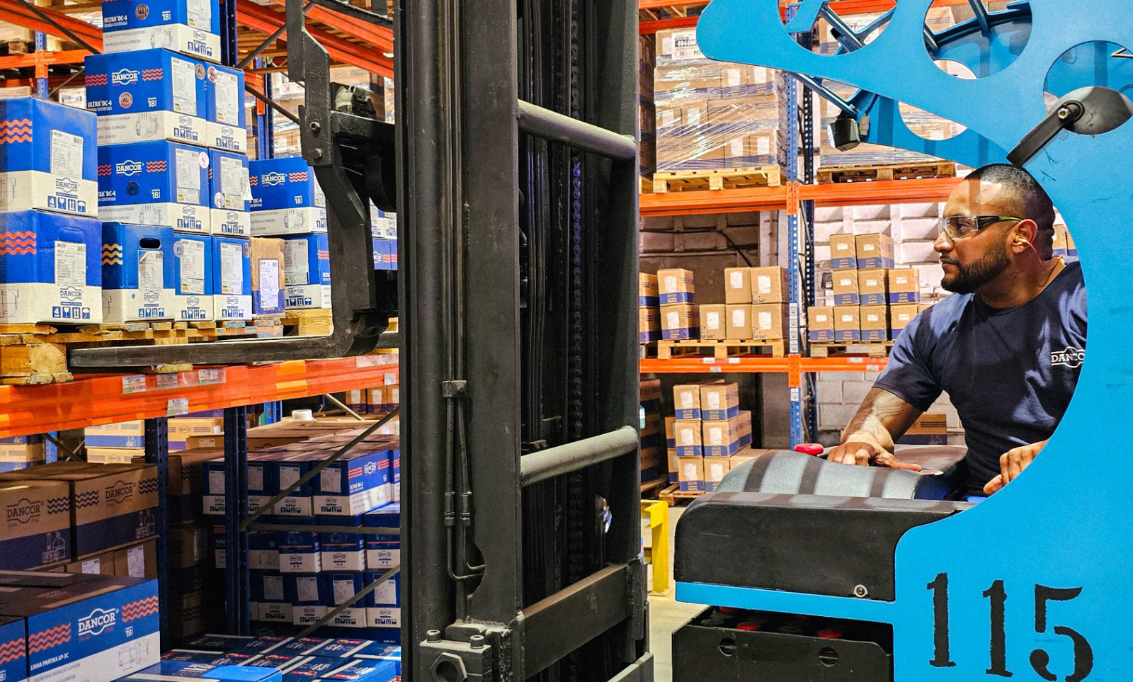 Operador de empilhadeira em um armazém logístico organizado, utilizando equipamentos de segurança e operando a máquina para movimentar pallets de caixas de produtos Dancor em prateleiras industriais de alta densidade.